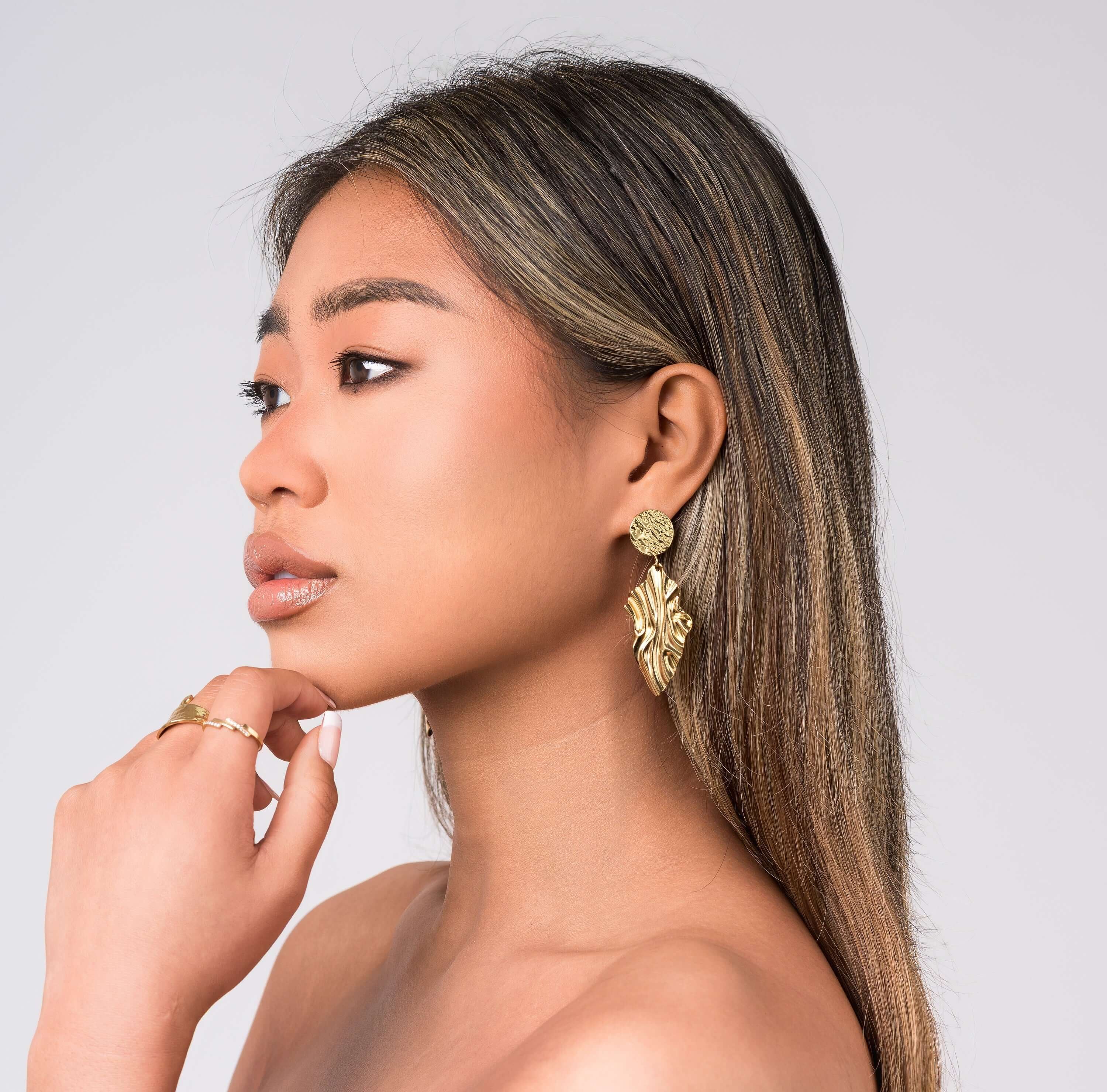 Model showcasing ARIIRA Layla textured drop statement earrings, featuring hammered discs and sculptural leaf design.