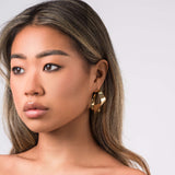 A model showcasing ARIIRA Alma Folded Ribbon Hoop Earrings, featuring a sculptural gold design, elegant and lightweight.