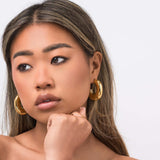 Stylish woman wearing ARIIRA Rhea Sculpted Dome Hoop Earrings, showcasing modern elegance in gold.