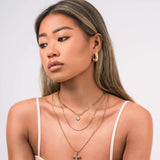 Model wearing ARIIRA Nour Layered Sun & Cross Necklace Set, showcasing layered gold chains and charms.