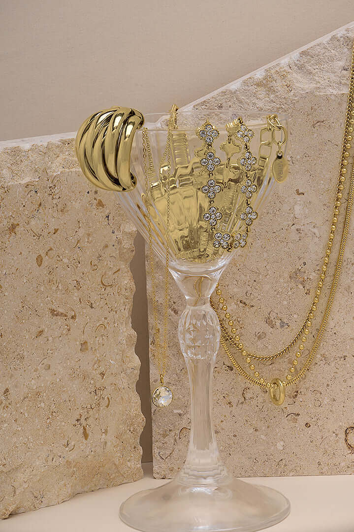 Elegant display of gold jewelry in a glass holder, featuring necklaces and rings, set against textured stone.