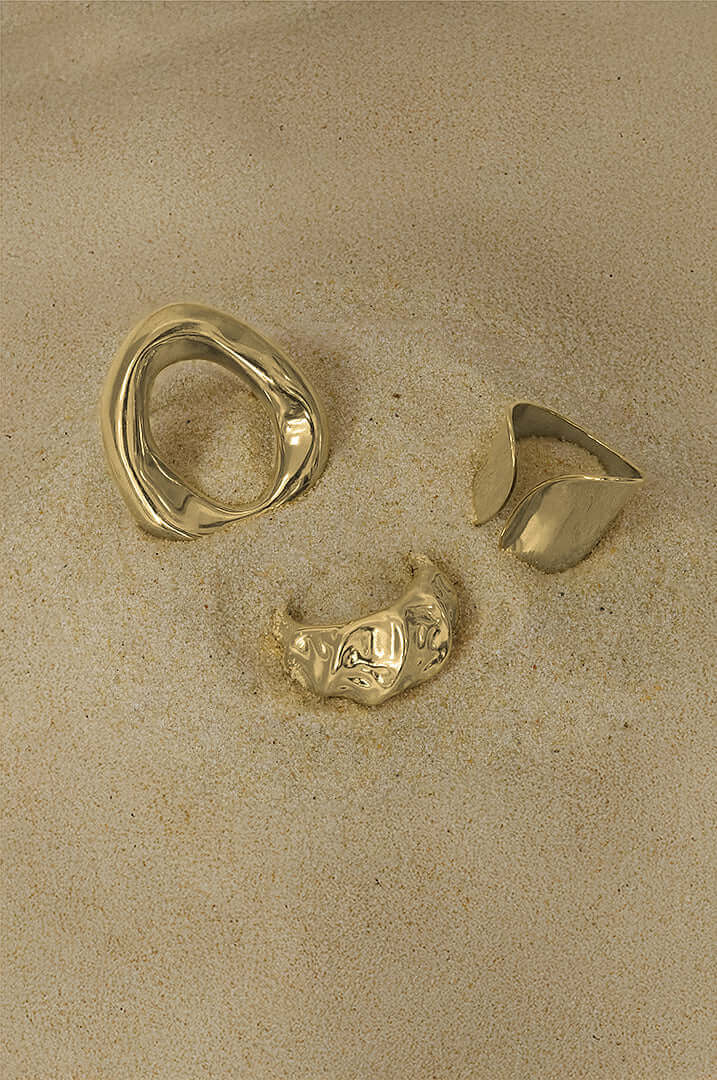 Three unique gold rings placed on a sandy surface, showcasing modern jewelry design and elegance.