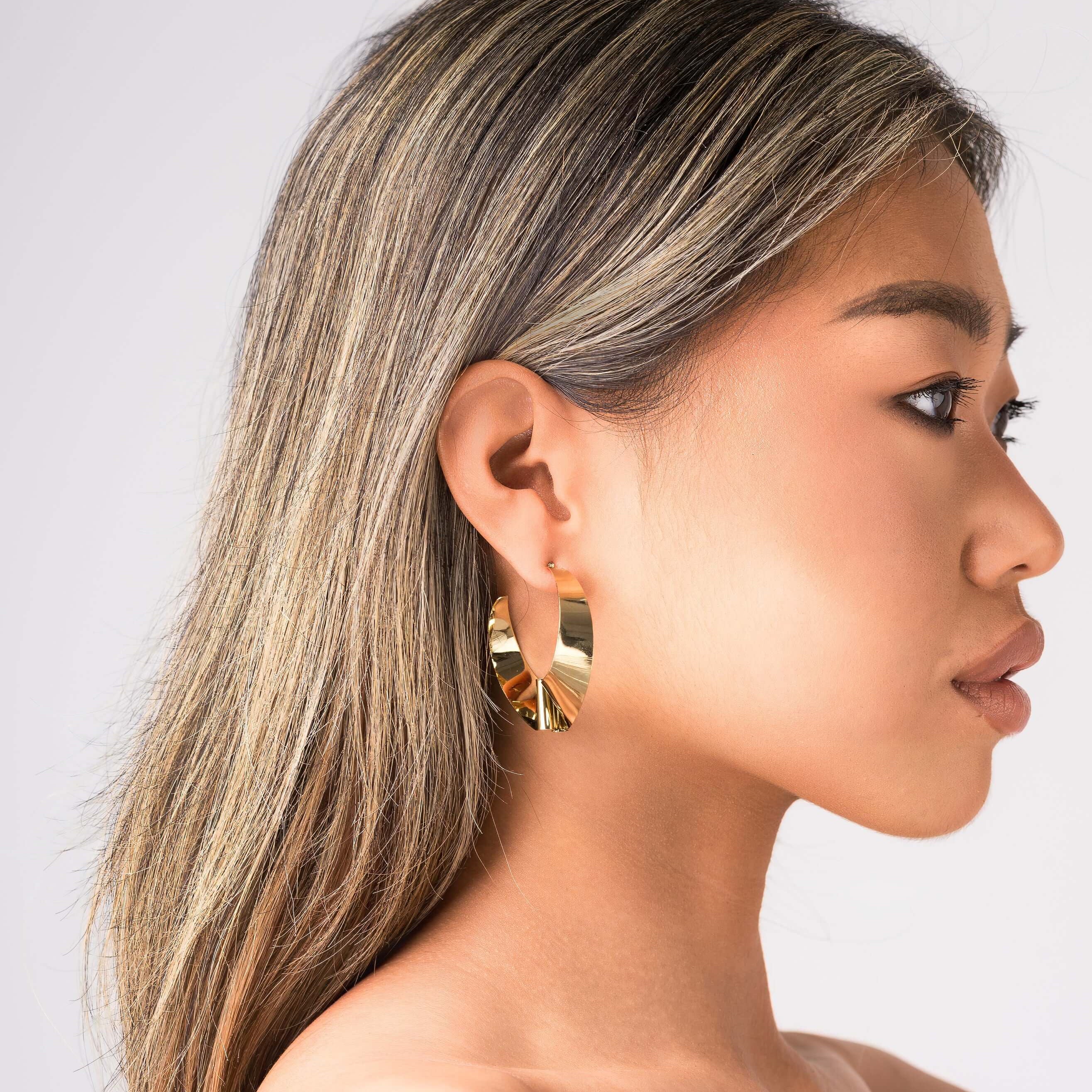 ARIIRA Alma Folded Ribbon Hoop Earrings in gold, showcasing their elegant design on a model's ear.