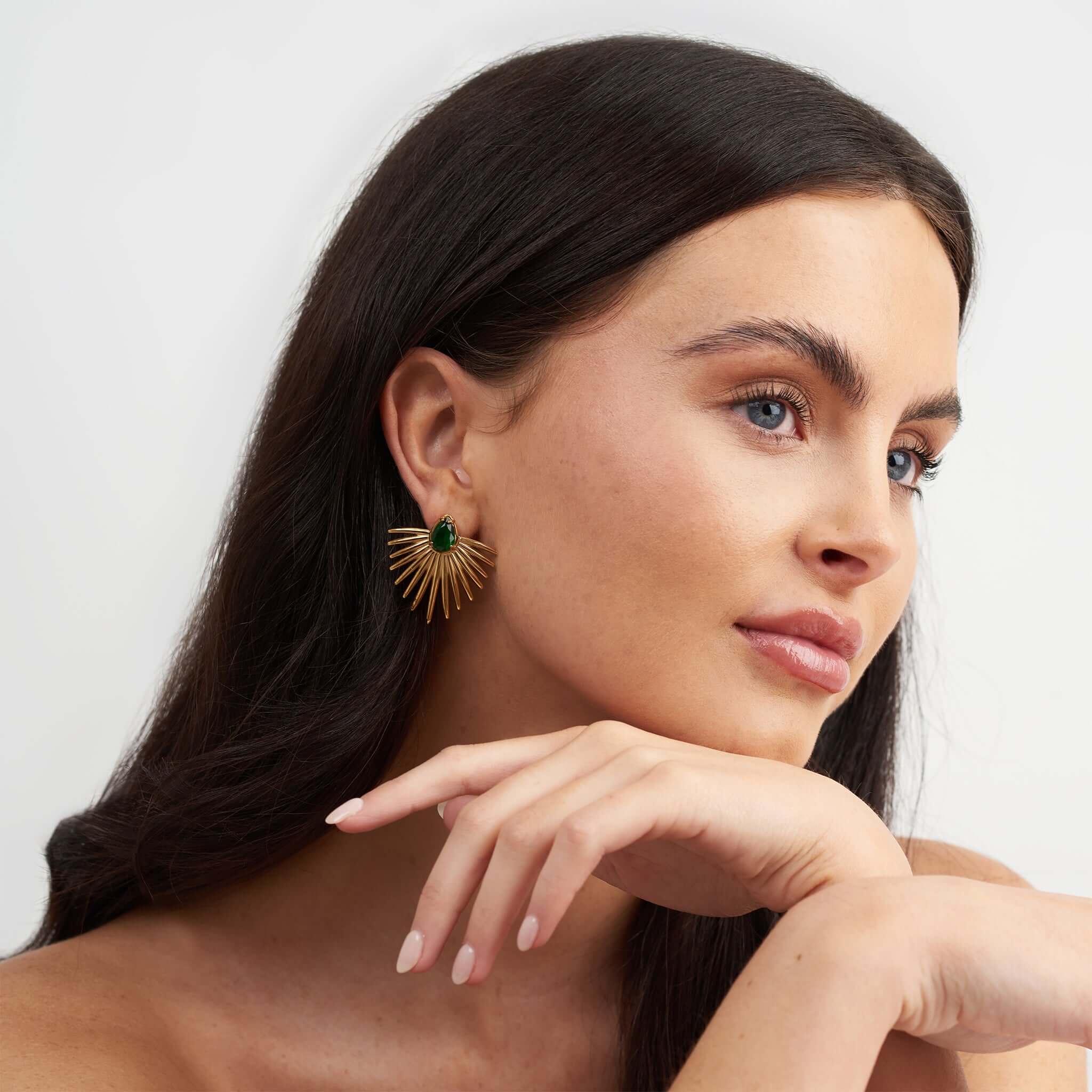 Elegant woman wearing ARIIRA Alina Emerald Radiance Fan Earrings with a green gemstone and golden rays.